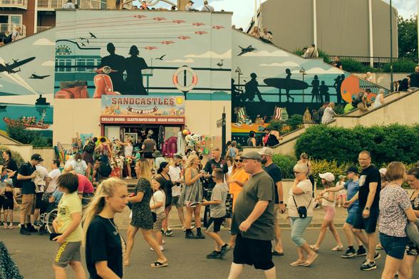 A bustling summer scene in Cleethorpes during Lancashire Armed Forces Day. Dozens of people walk across the street in front of a colourful seaside mural. The mural shows silhouettes of planes, soldiers, and surfers, along with bright beach imagery. In the middle is a gift shop called “Sandcastles,” its entrance crowded with visitors and racks of inflatable toys, hats, and beach goods. The atmosphere is lively, with families, children, and couples all moving in different directions, echoing the energy of a busy seaside festival.