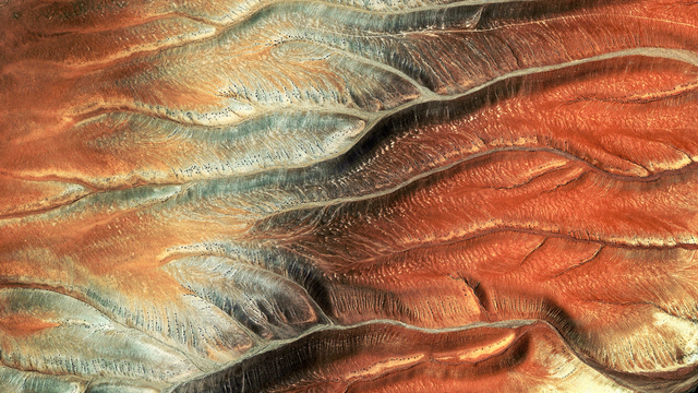 A satellite picture of a desert region showing eroded ridges and valleys. The terrain features layered sedimentary rock formations with colors ranging from red and orange to tan and gray.