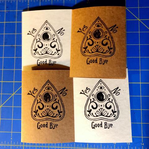 Four hand block printed cards, two on craft paper cardstock, and two on parchment pattered cardstock.
The print is of a ouija board planchette with curling filigree, a small triple moon over a flower at the bottom and an open eye towards the top point,  the viewing window is dark black. Around the outside of the planchette are the words “yes” on the left “no” on the right and “good bye” underneath