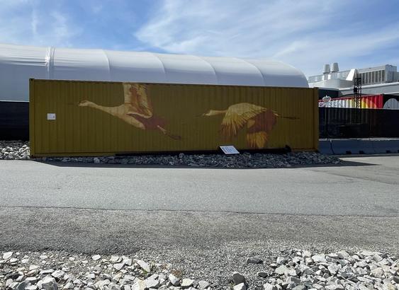 From the side of a paved road lined with gravel on each side, we look across to a tractor/trailor container sitting in the gravel on the other side, painted olive drab with two huge cranes in flight spanning the whole side of the container, painted in brown and tan tones. Behind the container is a fence. Along the other side of the fence is a white arched quonset building, and off to the side we can make out what must be the top of a bright mural in white, yellow, orange, and red.