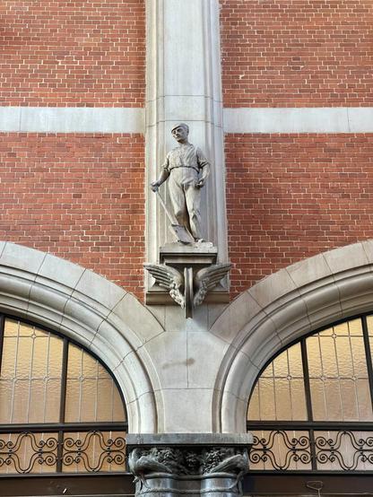 A lime stone statue of a train worker, with a shovel resting on the ground in his hand. The statue is on top of a building columns, between red bricks walls.