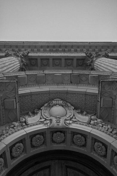 A low-angle, black and white photograph looking up at the ornate, classical-style archway of a grand building. The focus is on the intricate stonework, including a large, central cartouche with scrollwork, carved rosettes lining the arch, and the tops of two fluted columns with elaborate Corinthian capitals. The dramatic perspective emphasizes the architectural details and grandeur of the facade against a plain sky.