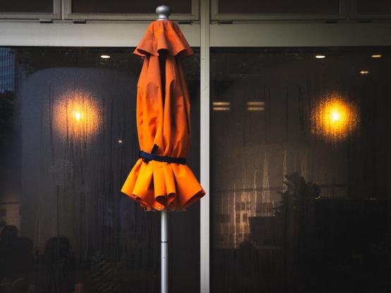 Folded orange patio umbrella with a blue strap against a window. The window has droplets indicating recent rain, and warm light shines from inside, casting soft reflections.