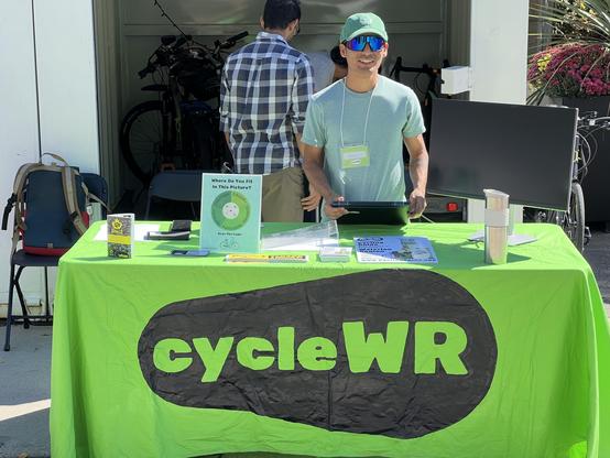 A table with a green table cloth that reads cycleWR. Behind the table someone is standing and looking at the camera.