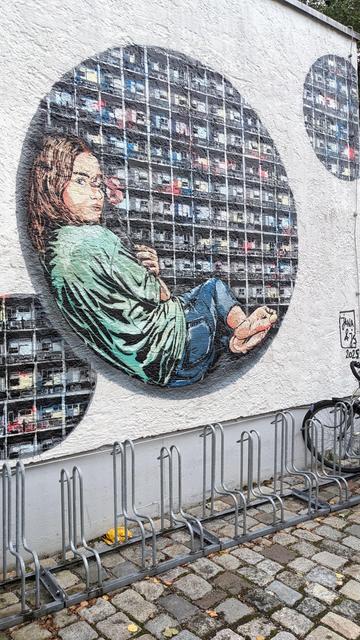 Behind a bicycle stand, a graffiti in stencil style is visible on the wall. It shows three circle depicting a grid like the facade of a house of same-size apartments. Each apartment facade has a different color. In the largest circle, a girl is seated along the round shape of the circle and holds a red flower in front of her left eye.
