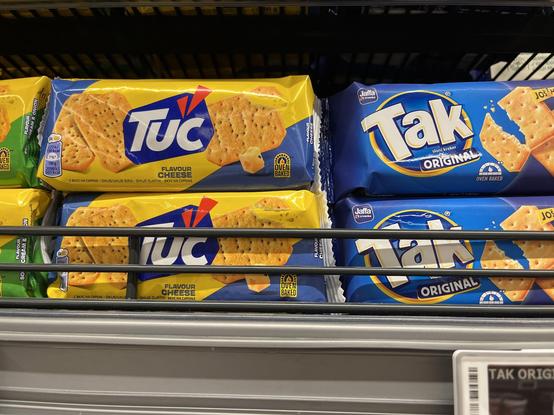 Shelf of Albanian grocery store with packages of seasoned crackers of the Tuc brand and saltines of the Tak brand right next to each other.