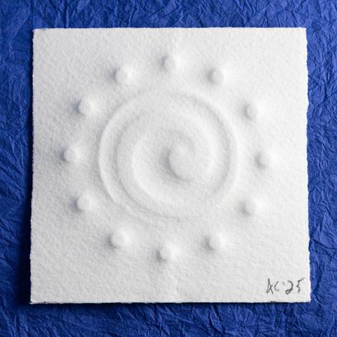An off-center counter-clockwise tapering spiral surrounded by twelve small circles is embossed on a small (70mm) square of toothy paper. It sits on a background of crinkled blue paper (momigami).