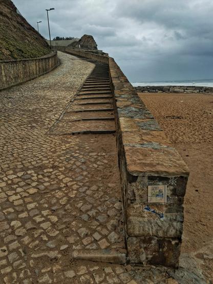 [pt] rua a subir em calçada à portuguesa, com um muro de pedra a separar da praia. Ao fundo o mar e o céu de um dia cinzento.

[en] Portuguese-style cobbled street, with a stone wall separating it from the beach. In the background, the sea and the sky on a gray day.
