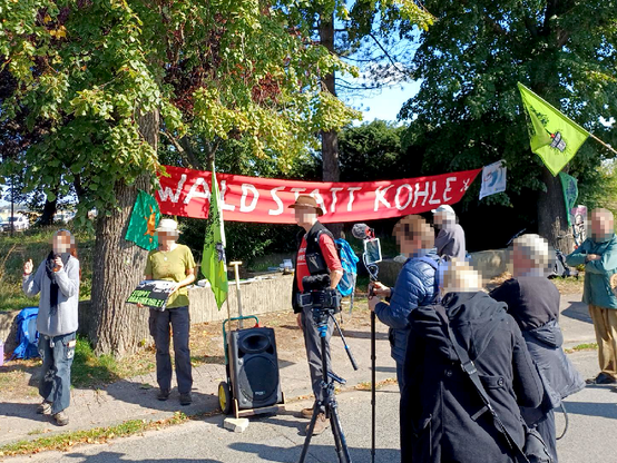 Unter einem roten Banner mit Aufschrift "Wald statt Kohle!" stehen Redner*innen, Kameraleute und Demonstrant*innen mit grünen Fahnen