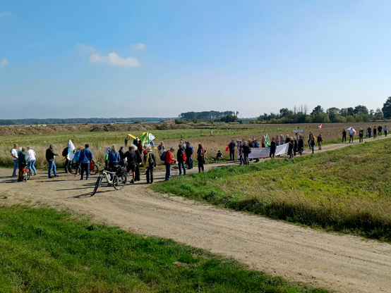 Dutzende Teilnehmer*innen des Spaziergangs stehen an der Wiese des BUND, im Hintergrund ein Erdwall von RWE und am Horizont das "Sündenwäldchen"