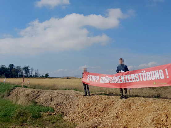 Auf einem Erdwall von RWE stehen Demonstrant*innen mit einem roten Banner "Stopp Grünflächenzerstörung!", im Hintergrund der Hambacher Wald
