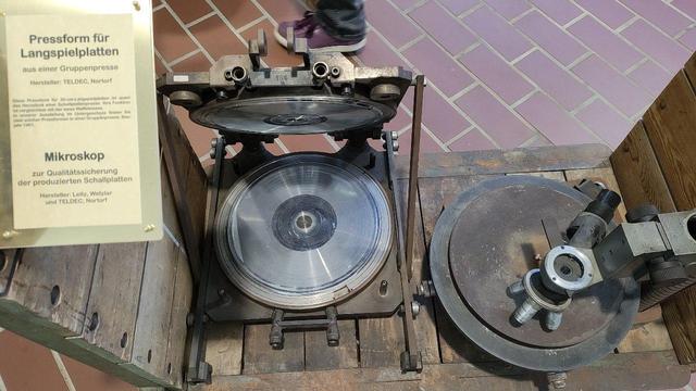 Zwei alte Maschinenteile aus dem Betrieb.
Links eine Pressform für Langspielplatten. Sie sieht im Prinzip aus wie ein Waffeleisen.
Rechts ein Mikroskop, das auf einem metallenen Plattenteller montiert ist. Es diente zur Qualitätssicherung.