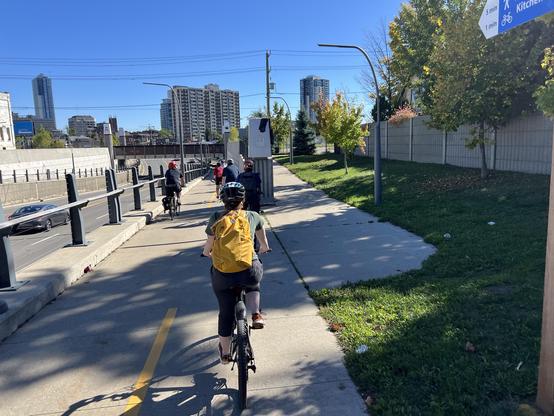 A multi-use path in an urban setting recedes into the distance, passing under a railroad bridge. In The foreground and middle ground, many cyclists are riding along the path. The sky is clear and blue.