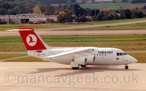 Side view of a high-winged, 4 engined jet airliner taxiing from left to right.
The plane is almost entirely white, with blue "Turkish" titles on the upper forward fuselage, just in front of the wings.
There is a small Turkish flag under the cockpit windows, and the blue name "Usak" on the nosecone, just above the nosewheel.
The black registration "TC-THJ" is on the rear fuselage, under the tail.
The tail is red, with a white circle containing a stylised flying bird.
Grey concrete apron fills the foreground, with large grassed areas lining a grey runway and taxiway in the background, and buildings nestled in trees in the distance.