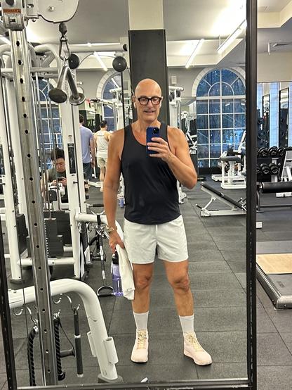 Me with a shaved head,  wearing a black tank top, light shorts, and pink shoes while holding a water bottle and a towel. They are taking a selfie in a gym mirror, with various gym equipment and other gym-goers visible in the back.