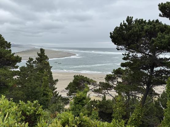 Facing south from a ridge high above the north shore overlooking the mouth of the Siletz River, two small fishing boats are headed west toward the sea. The sky us filled with gray clouds the seas are dark greenish gray. The Salishan spit is seen on the south side of the river and the bay is seen near the top right. Large conifers fill the right quarter of the image and the bottom third of the image.