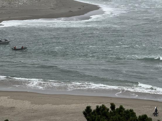 Facing south from a ridge high above the north shore of the Siletz River, two small boats are seen carrying four fishermen towards the sea. The Salishan spit is across the river with a few logs and/or sea lions resting near the water.