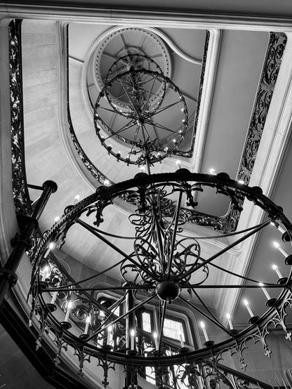 Black and white image looking up a multiple-level stairwell that has three large iron rings that hold candles. The iron work's basic structure is like a wagon wheel, but it has a lot of embellishment. The ironwork of the stair rails is also elaborate. Biltmore House in Asheville, North Carolina. It is the largest privately owned houses in the US.