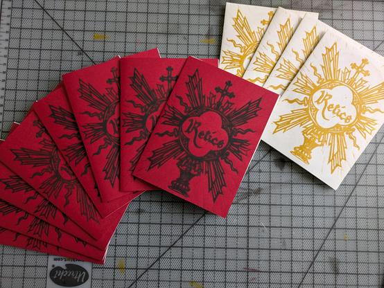 Several of my relics lino zine fanned out on a gray cutting mat. Most of them have the red cardstock cover, but a few of them are coverless, so you just see the gold ink print on the white interior paper.