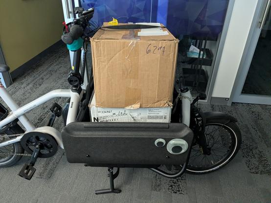 The Cargo Demon's frunk folded open with two shirt boxes stacked inside. The second box protrudes all the way over the top edge of the frunk and is held in place by a spare Bakkie cinch and optimism. (It was only going half a mile, it was fine.)