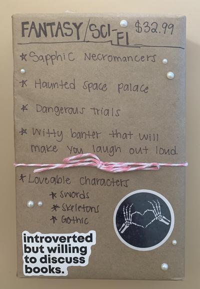Photo of a mystery book, wrapped in brown paper. The book is tied with pink-and-white string, and has a sticker of two skeletal hands making a heart sign, and a sticker that reads “introverted but willing to discuss books.”. Handwriting on the paper reads:
“FANTASY/SCI-FI
$32.99
•Sapphic Necromancers
•Haunted Space Palace
•Dangerous Trials
•Witty banter that will make you laugh out loud
•Loveable characters
•Swords
•Skeletons
•Gothic”