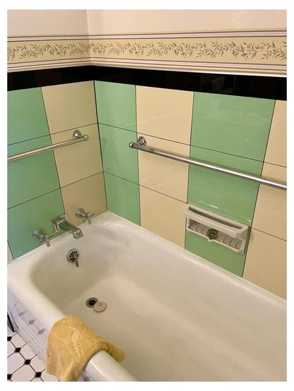 Vertical photo of an interior bathroom showing a bathtub with pastel green and beige tiles on the walls. The tub features a yellow washcloth hanging over the side, two silver wall-mounted grab bars, and plumbing fixtures visible at the far end. A decorative tile border with a floral pattern is present at the top of the wall. The floor is visible, composed of white and black hexagonal tiles.