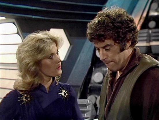 Claude 3.7 describes the image as: "This image shows a scene set in what appears to be a spacecraft interior with distinctive futuristic design elements typical of classic science fiction television from the late 1970s/early 1980s. 

The scene features two individuals in conversation - a person with blonde feathered hair wearing a navy blue outfit with decorative gold accents on the shoulders, and another person with curly dark hair wearing a brown jacket over darker clothing.

The spacecraft set has hexagonal doorways and technical panels visible in the background, creating the atmosphere of a command center or bridge. The lighting and production style have the characteristic look of BBC productions from that era, with the somewhat confined set design and specific costume aesthetics that were common in science fiction television productions working with limited budgets."

Blakes7Bot, version 3.9.33.