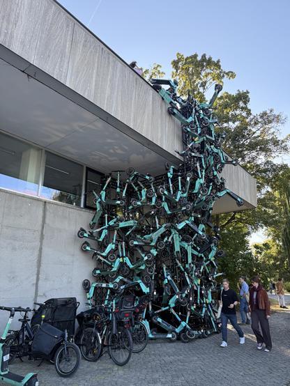Installation of many e-scooters, hanging on a wall outside a concrete wall. Some people walking by, in the background, some trees, and the sky is blue