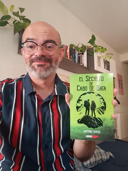 Calvo barbudo muestra libro de portada verde y negra en habitación