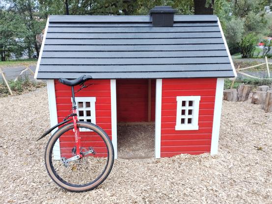 A small red playhouse with white trim and a black roof sits on a bed of wood chips. Its open doorway and two white-framed windows suggest a child-sized structure. A red unicycle leans against the front. In the background are trees and a paved path.
