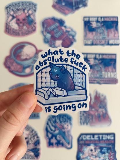 Hand holding up a sticker of a kitty that woke up in psych ward, called "what the absolute f*ck is going on"