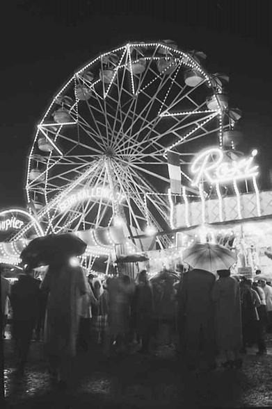 Treiben auf dem Volksfest: Im Vordergrund MEnschen mit Schirmen, dahinter Stände, ganz im Hintergrund das Riesenrad.
