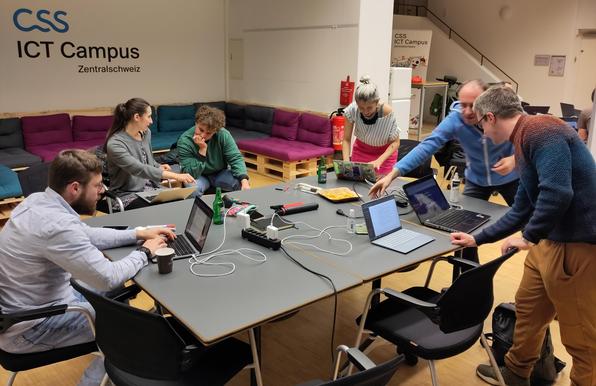 Ein Foto eines vergangenen Hackergarten Events mit sechs Personen rund um einen Tisch, teilweise sitzend, teilweise stehend, fast alle mit Notebook und miteinander am Diskutieren. Im Hintergrund sind noch weitere Personen mit Notebooks zu sehen.