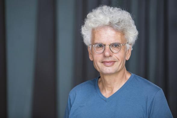 Porträtfoto (Torso) von Roland Stimpel. Er trägt ein hellblaues T-Shirt, steht vor einem dunklen, graublauen Vorhang und blickt direkt in die Kamera.