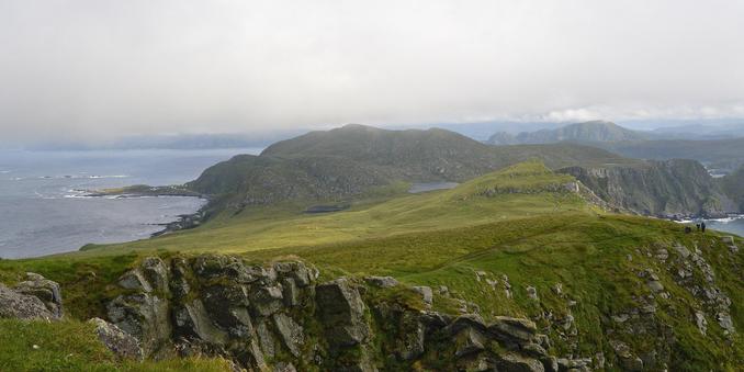 A photo of a green hilly landscape with jagged coasts. There is a rocky cliff in the foreground. The sky is cloudy.