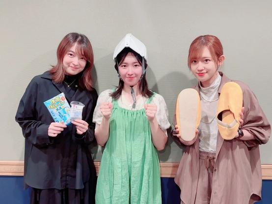 Three women are standing together indoors, smiling at the camera. The woman on the left is holding a package and a small item, the woman in the center wears a white helmet-like device and a green dress, while the woman on the right is holding a pair of yellow slippers.