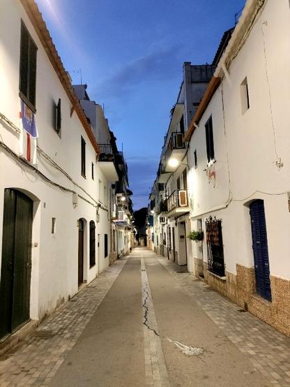 Blick ein eine Straße in Sitges. Es ist früher Morgen, die Straße ist menschleer. Sie verläuft pfeilgerade zum Horizont, recht und links mit älteren Häuser gesäumt, einige beflaggt mit Regionalflaggen - ein fast symmetrisches Bild. Am Himmel kündigt sich das Morgenrot an.