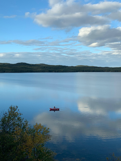 Hunter, my neighbor’s son, sits serenely in his small red boat. The water, smooth as glass, reflects the sky above, creating the illusion that he is paddling directly into a cloud.