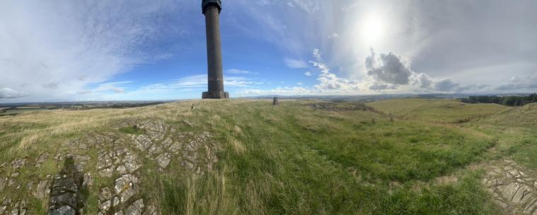 A panoramic view off the memorial with under a blue sky with white clouds, the sun peeking from behind one, a dug in front of the memorial and the trig point to the right. The hills in the background mark the English border
