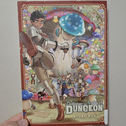 Photograph of a white person's hand holding Delicious in Dungeon, Volume 8 against a light colored wall. The cover is bordered in red and gold with the title in the lower right-hand corner. To the left side of the cover is a dark skinned, blue eyed character in plate mail armor kicking a blue mushroom creature. Behind them are a group of other adventurers and a ton of various sized living mushroom creatures.