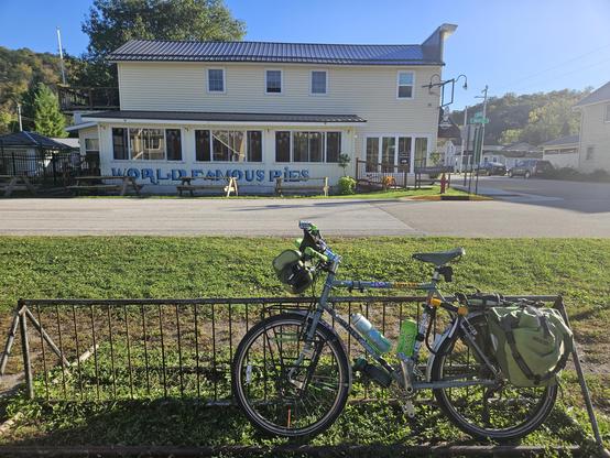 A bicycle with green bags leans against a brown and grey bicycle parking rack, green grass in front of a grey asphalt street. Under a blue sky, an almost yellow 2 story building is in the background. In blue and black stylized letters on the wall are the words "World Famous Pies".