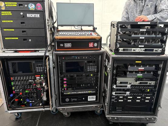 Mehrere große Flightcases voller Equipment stehen im Seitenbereich einer Bühne und werden gerade verkabelt.

Links ein Rack mit großem analogen Mikrofon Splitter und Behringer WING Rack Mischpult, dadrüber ein Case mit Schubladen voller Funk-Equipment.

In der Mitte ein Rack mit SPL Track One Channel Strip, Shure ULXD4D Funkempfänger, Shure PA421A Antennencombiner, Shure PSM1000 IEM-Funksendern, Furman Esoterik-Steckerleiste, Behringer WING Rack Mischpult, Swissonic 19" Plasterouter, obendrauf ein Mischpult-Controller bestehend aus Waves FIT Controller, HannsG HT225 Touchscreen und unbekanntem Windows-PC mit MixingStation.

Rechts ein Rack mit einer dicken USV, 19" AA-Akkuladegeräten 2x iConnectivity mioXL RTP-MIDI Konvertern, und einer Wagenladung Neural DSP Quad Cortex und Sennheiser ew G4 Funkempfängern (Gitarren) welche sich in das kleine Rack obendrüber fortsetzt.

Hinter dem rechten Rack steht eine Person in grauem Pullover die gerade verkabelt.