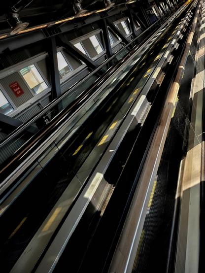 A tilted photo of a set of parallel moving walkways inside an airport tunnel, with yellow directional markings and windows along the left side.
