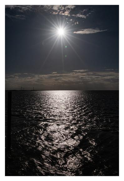 Foto im Hochformat. Blick über das Meer. Der Horizont ist genau in der Mitte des Bildes. Der Himmel ist leicht bewölkt, oben in der Mitte ist die Sonne. Von ihr gehen viele Sonnenstrahlen ab. Das Bild ist allgemein dunkel gehalten, das dunkle Wasser sieht fast aus wie eine feste Fläche aus dunklem Matsch.