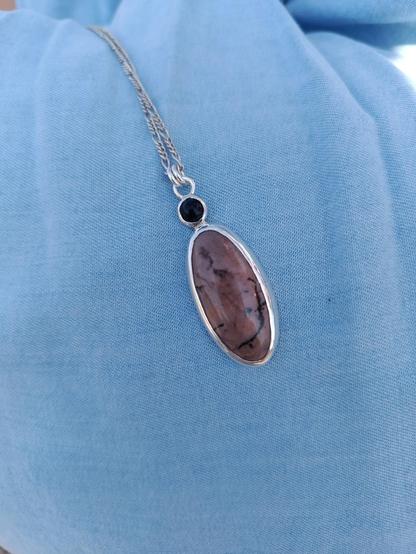 a pendant with silver chain. in has two gemstones one small round black colored and a bigger one oval shaped pink with black lines. it's placed on blue textile