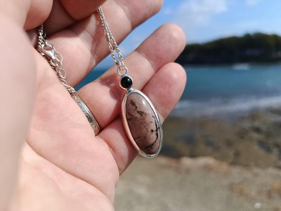 a pendant with silver chain. in has two gemstones one small round black colored and a bigger one oval shaped pink with black lines. it's hold by a hand, in the background in a distance tm there is a shore