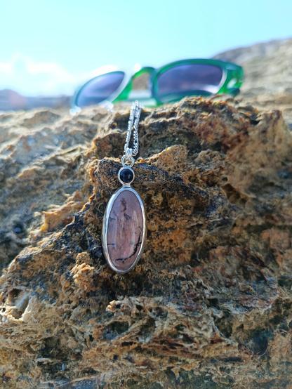 a pendant with silver chain. in has two gemstones one small round black colored and a bigger one oval shaped pink with black lines. it's placed on a brown rock
