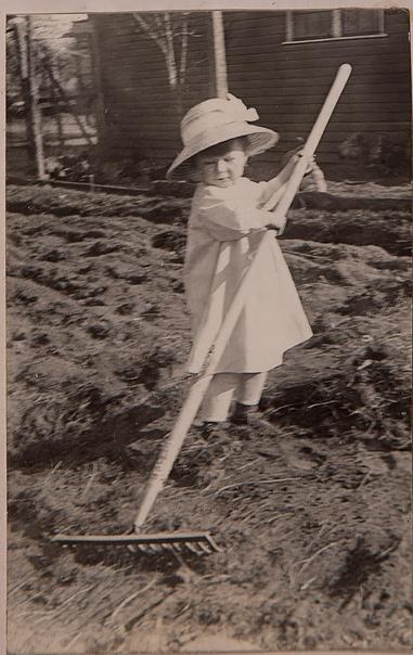 Little girl in a garden with a large hoe - she is wearing a white dress and white hat