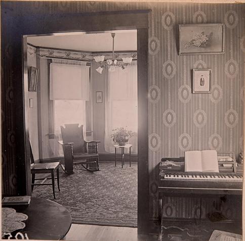 Interior photo of a family home - room closest to the camera has a piano, and striped wall paper. The other room has rocking chair and a side chair, big bay windows with light streaming in