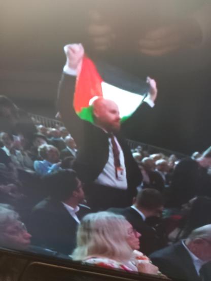 Man standing amongst seated people. He has the flag of Palestine above his head